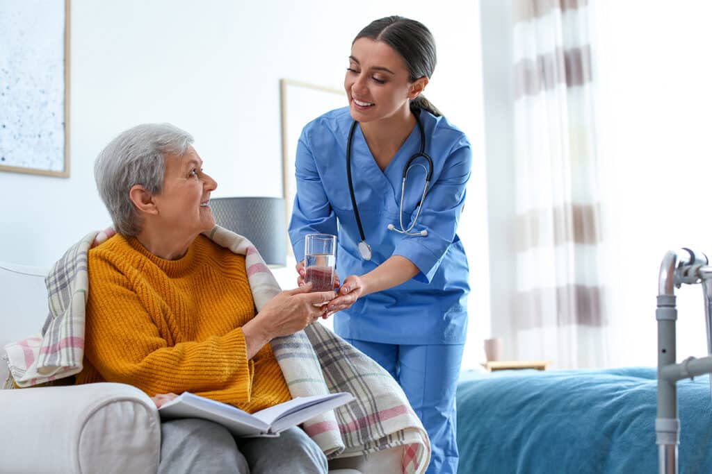 Smiling nurse in blue scrubs helps an elderly woman in a yellow sweater drink water beside a bed.