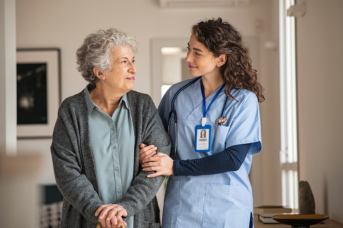 Nurse in blue scrubs holds arm of older patient in gray sweater as they walk
