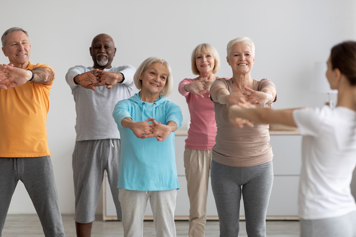 Older adults follow a fitness instructor in a gentle group stretching class, extending clasped hands forward