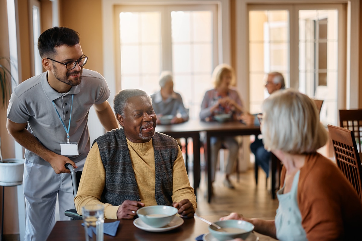 Caregiver assists a smiling older man at a community café table, while an older woman joins him
