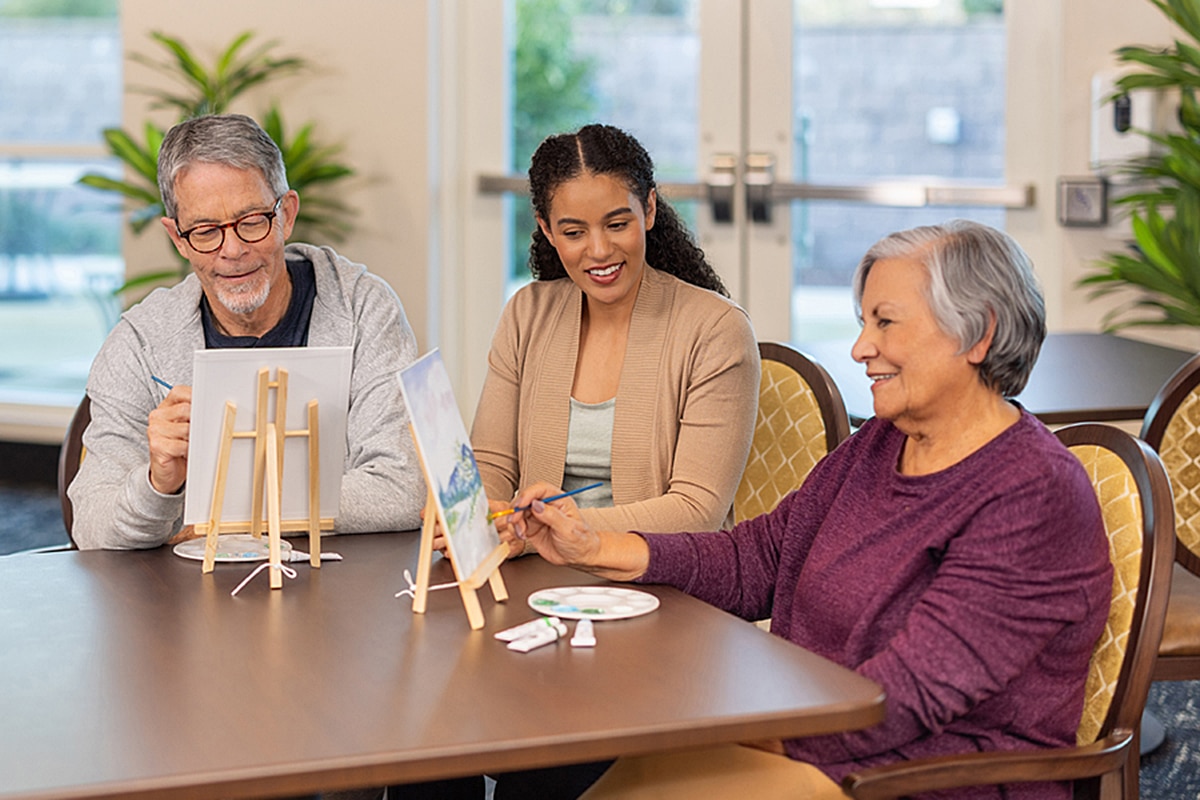 Three adults enjoy a cheerful painting activity together at a table, using easels and palettes in a bright, welcoming room.