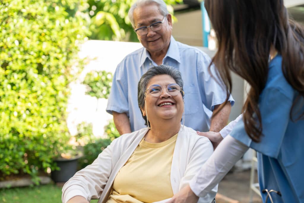 Smiling older woman in glasses sits outdoors as a caregiver and older man stand beside her