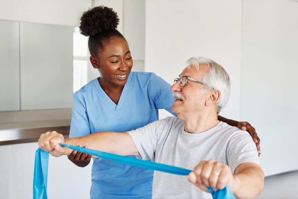 A healthcare professional in blue scrubs assists an older man with white hair and glasses as he stretches his arms using a blue resistance band.