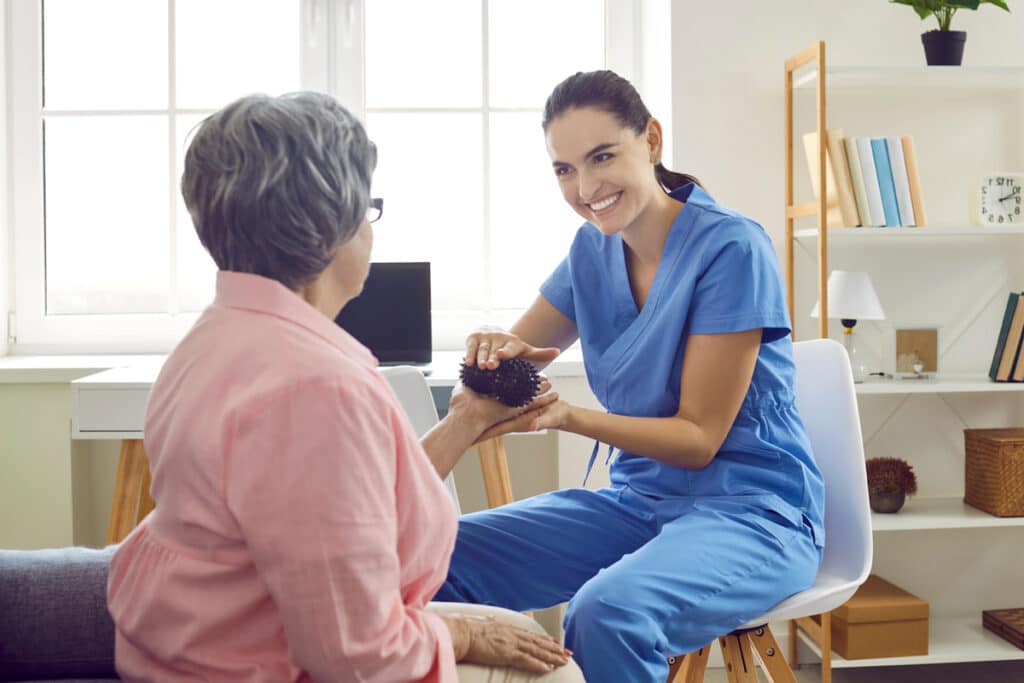 A healthcare professional in blue scrubs assists an older woman with a hand exercise using a textured therapy ball.