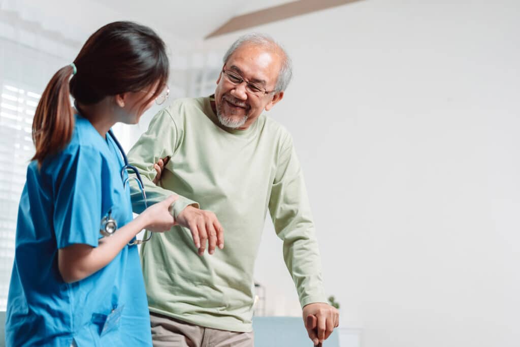Smiling nurse in blue scrubs supports an older man with a cane during a health check