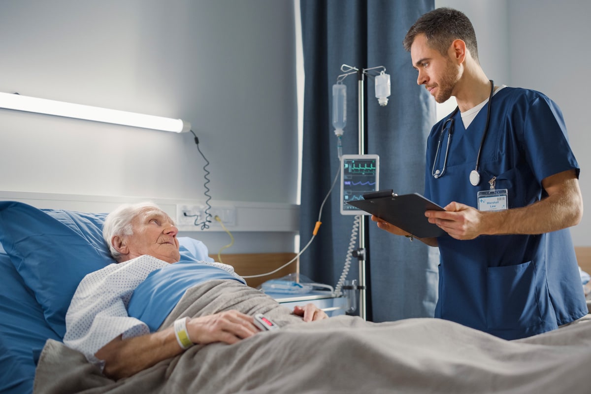 An older patient rests in a hospital bed while a healthcare professional in blue scrubs reviews information on a tablet beside them.