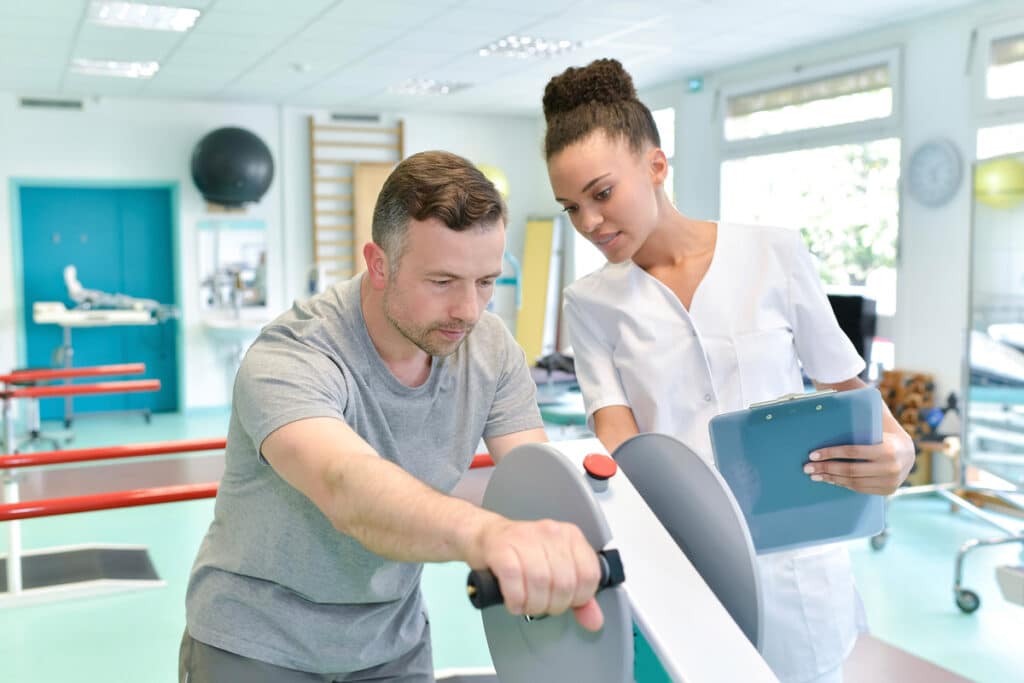 A man uses a rehabilitation exercise machine while a healthcare professional observes and takes notes in a bright physical therapy clinic.