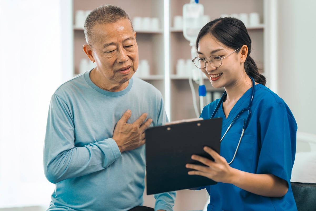 Nurse in blue scrubs reviews clipboard with patient in blue shirt with hand over his heart