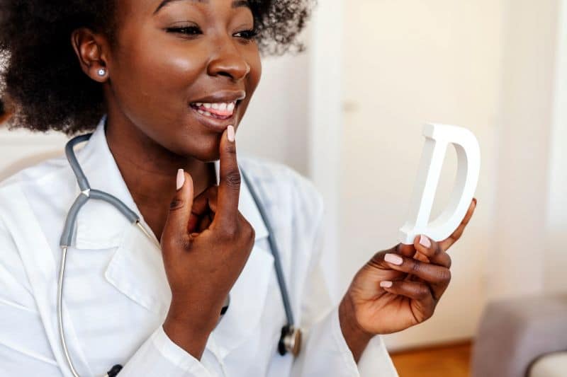 Nurse in white lab coat pointing to lip while holding the letter D