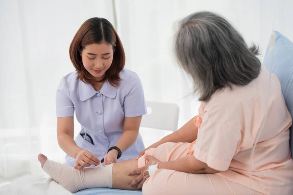 Healthcare worker gently examines an older woman’s bandaged leg at home, offering attentive care in a bright, calm setting.